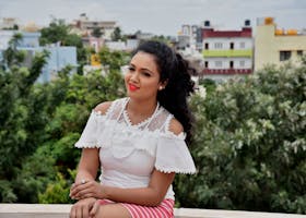 A cheerful woman sitting outdoors in a white lace top against a cityscape backdrop.